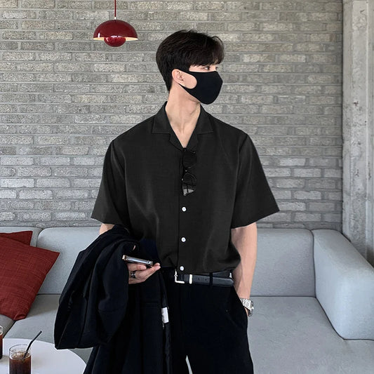 A man in Korean streetwear—a black mask, Baesic World Korean Shirts short-sleeve shirt, and black pants—stands indoors by a gray sofa, holding a jacket and phone. A drink and red pillow are on the nearby table and couch.